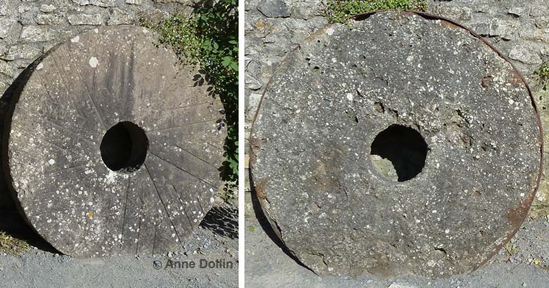 Two old millstones standing against a wall at Carew Castle in Wales, UK. One is made from a fine-grained sandstone while the other is a conglomerate containing rounded white pebbles.